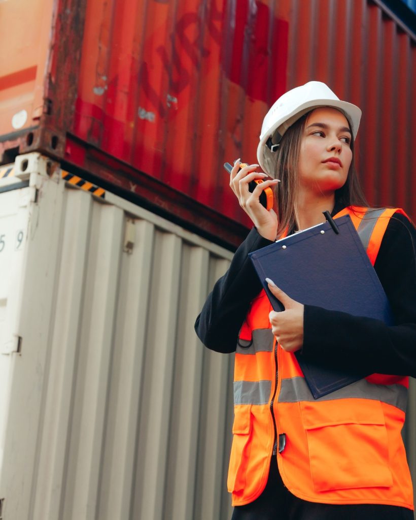 Portrait of engineer woman with walkie-talkie in white helmet and vest in container terminal